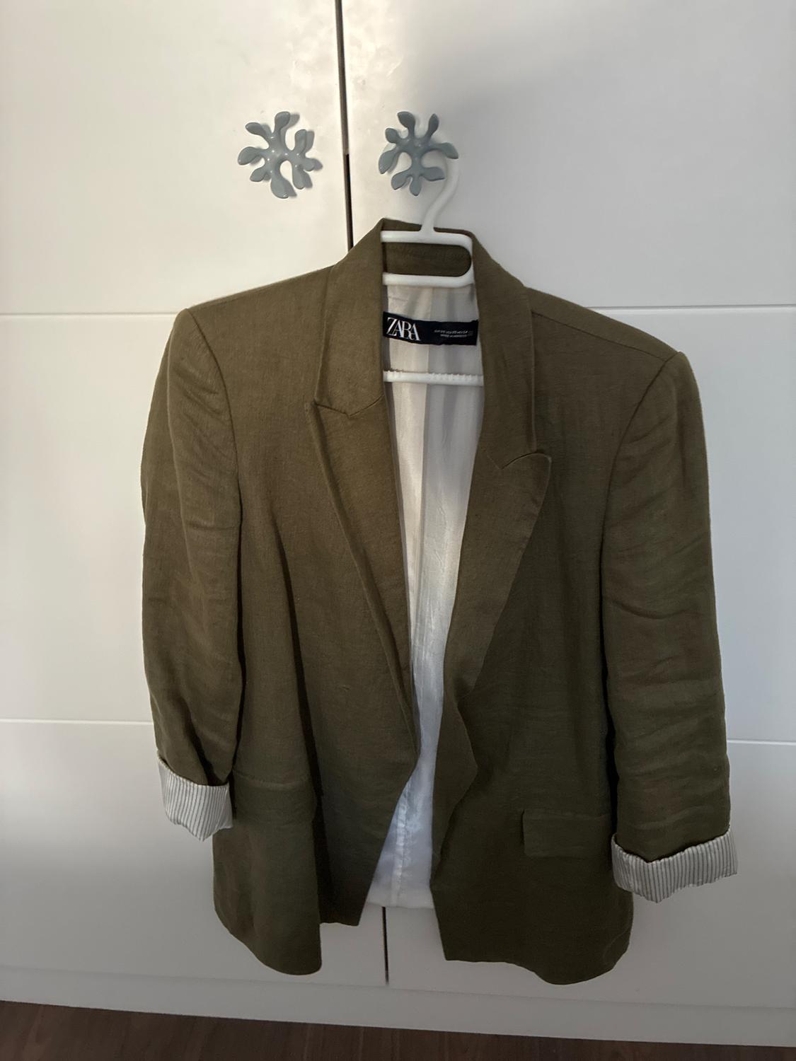 Olive green jacket hanging in a white wardrobe. Sleeves rolled up showing a striped lining. Black label with the text ZARA.
