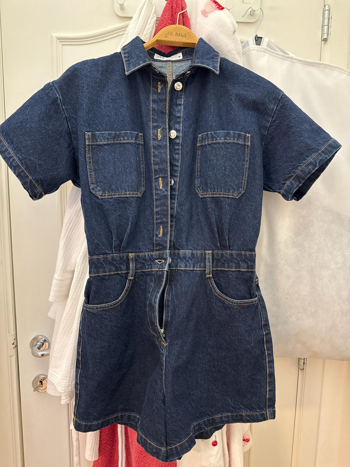 Dark blue denim playsuit on a hanger with J.C. RAGS text and a Zara label, hanging on a door. It has short sleeves and pockets.