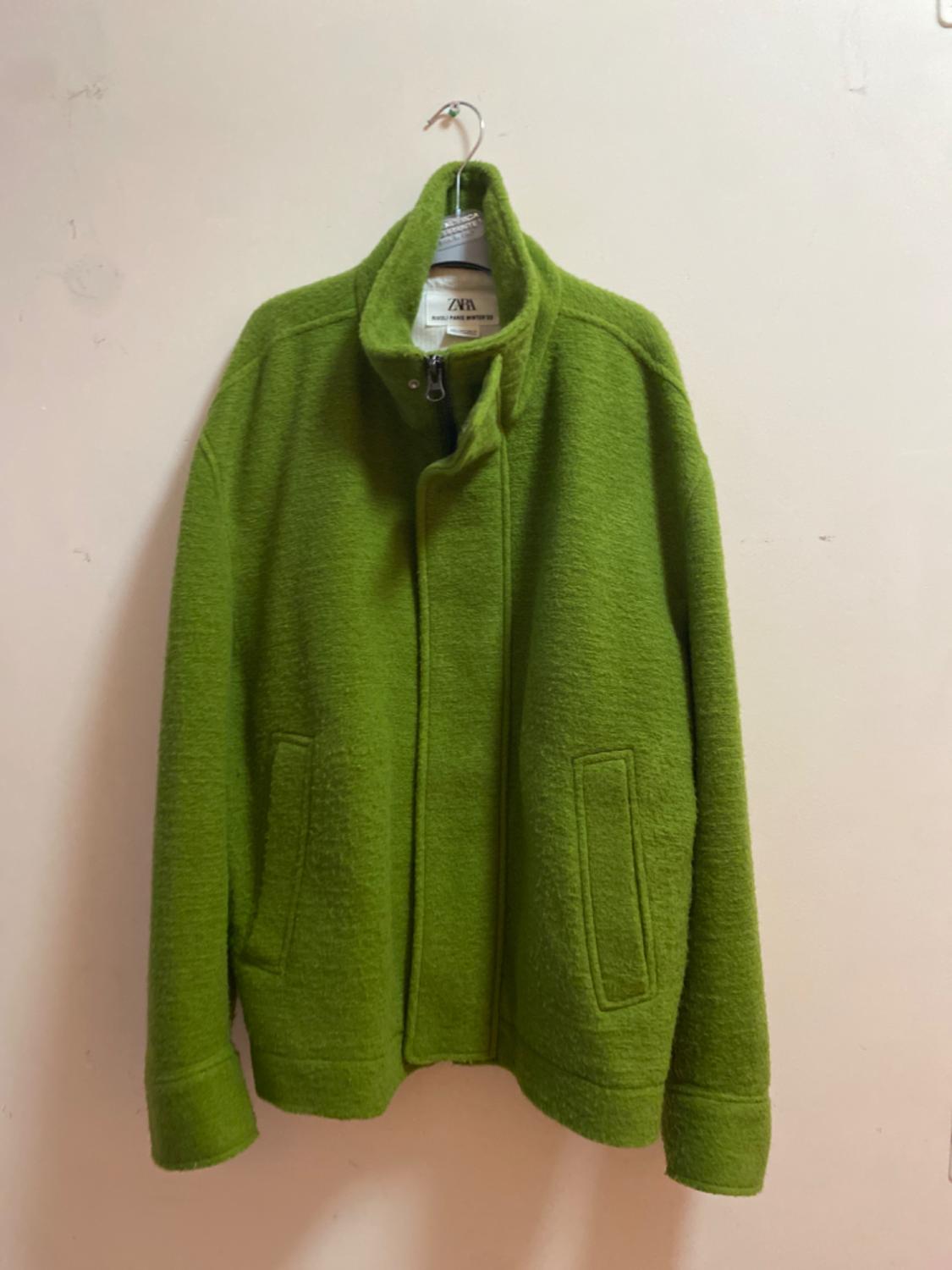 Green jacket hanging on a hanger against a beige wall. The inner label says "ZARA".