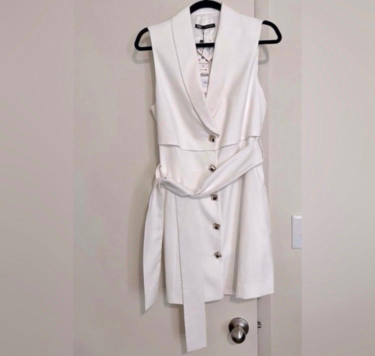 Sleeveless white dress with buttons and belt, hanging on a black hanger over a beige door. Label visible on the collar.