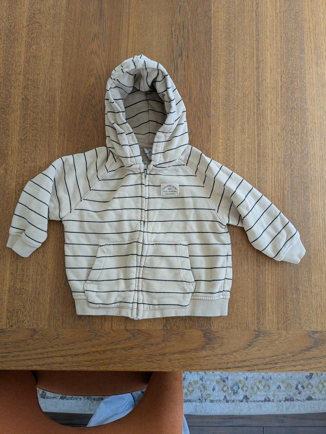 Kids' hoodie with black stripes on a beige background, placed on a wooden table. Visible tag on the chest.