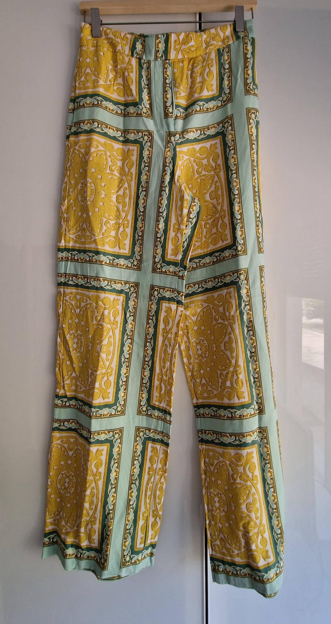 Long trousers with an ornamental panel print in shades of yellow and green on a light background, hanging on a wooden hanger.