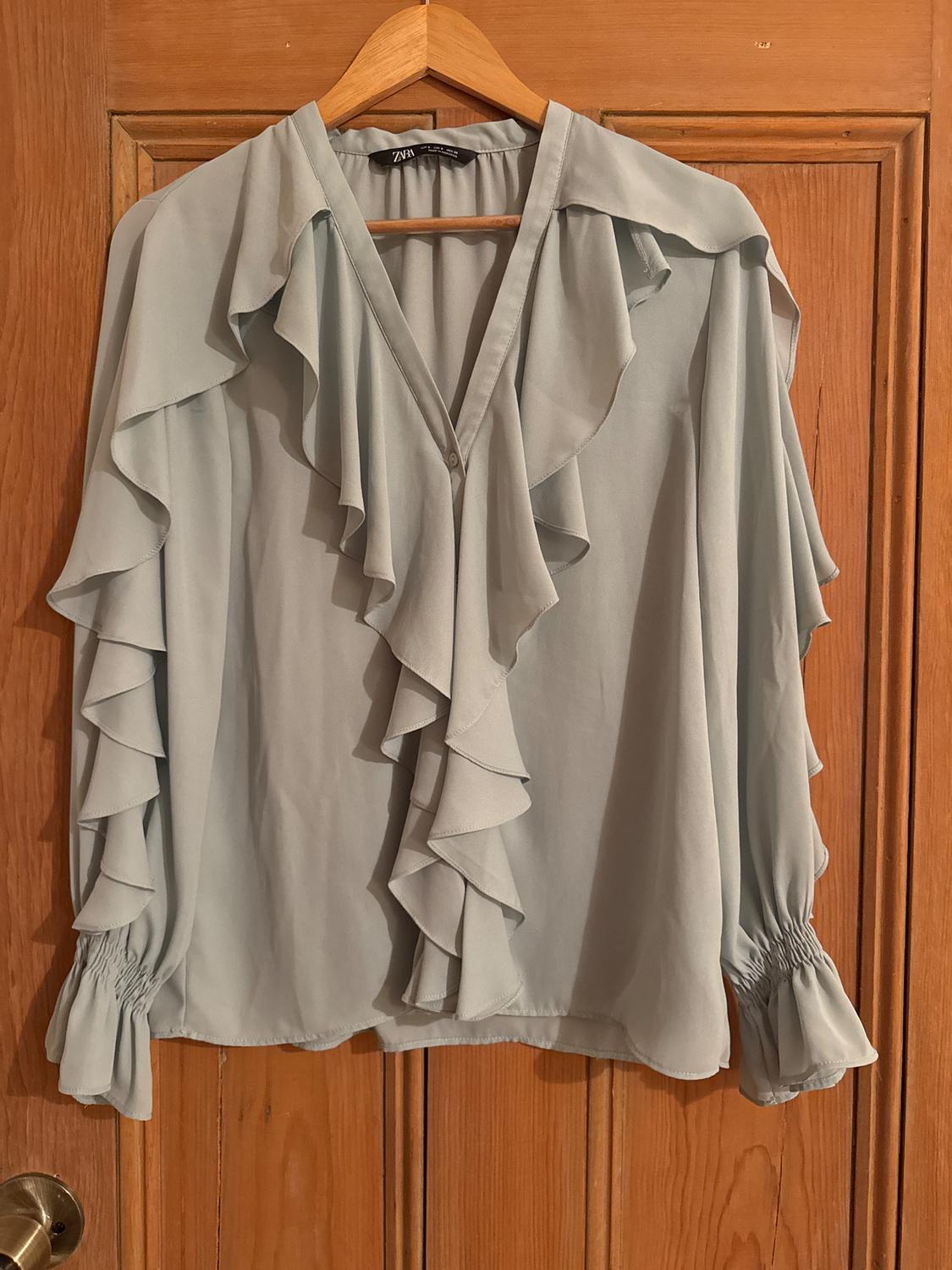 Light blue chiffon blouse with ruffles on the front and long sleeves with elastic cuffs, hanging on a wooden door.