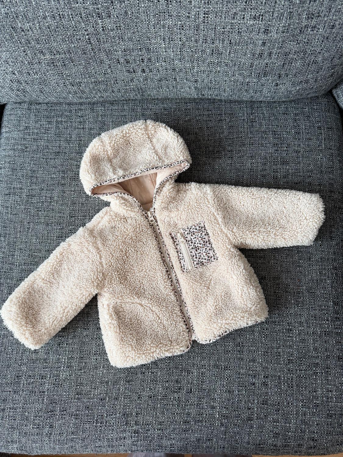 Baby jacket with soft fabric hood and zips, on a grey sofa. Decorative pocket with leopard print.