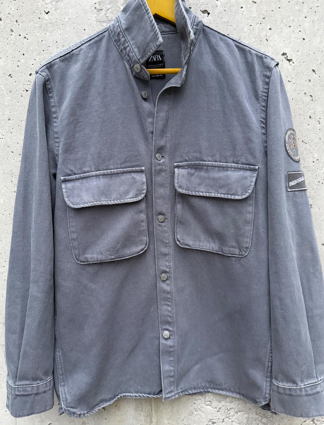 Grey long-sleeved shirt with two front pockets and a high collar, hanging on a yellow hanger against a white wall background.