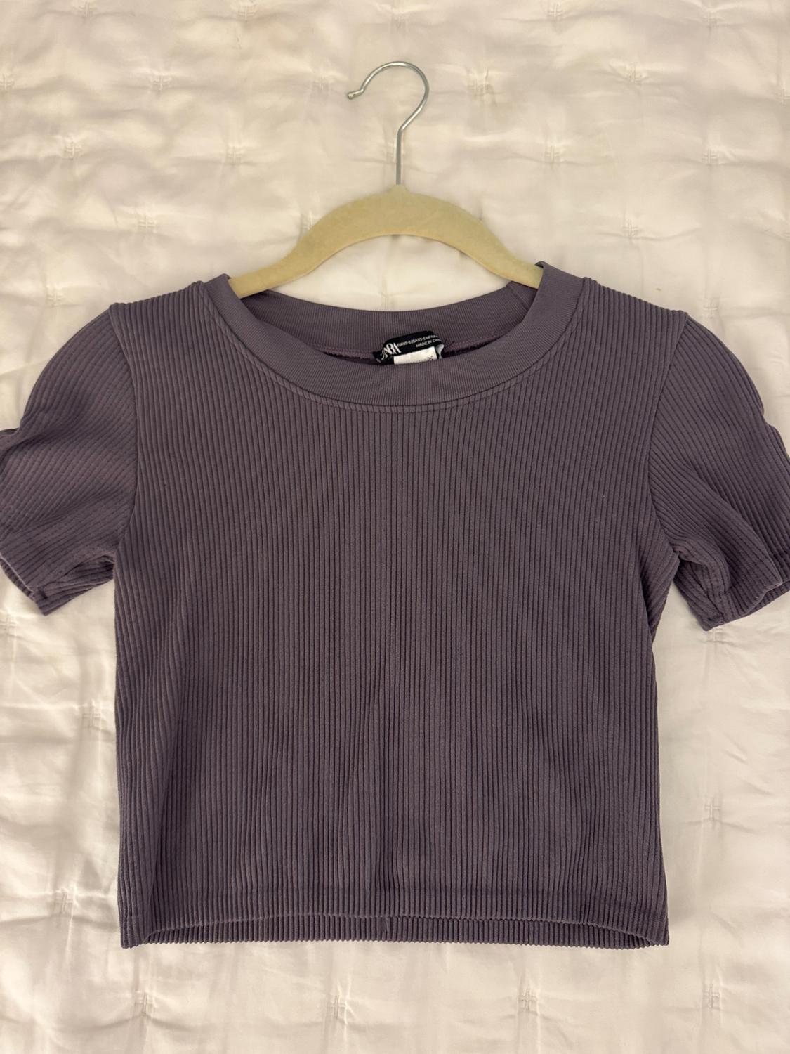 Ribbed crop top in lilac, featuring a round neck and short sleeves, hanging on a hanger over a white quilted bedspread.