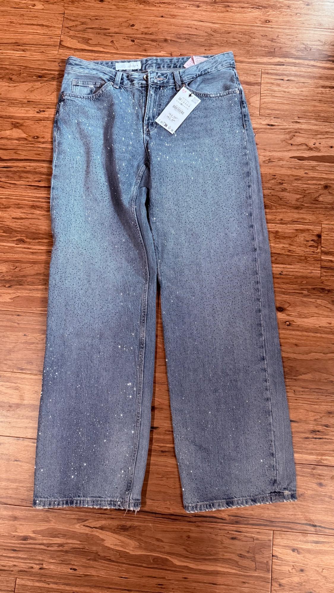 Light blue wide-leg jeans with all-over glitter, laid out on a wooden floor. They still have the tags on.