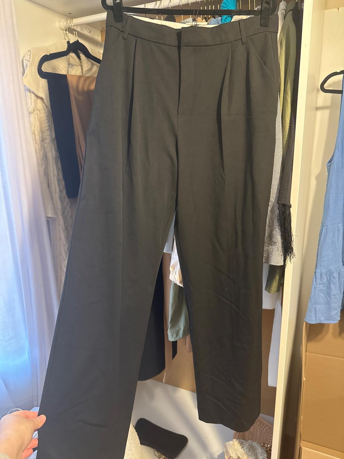 Dark trousers are hanging in a wardrobe, surrounded by various items of clothing. A hand is holding the left trouser leg.