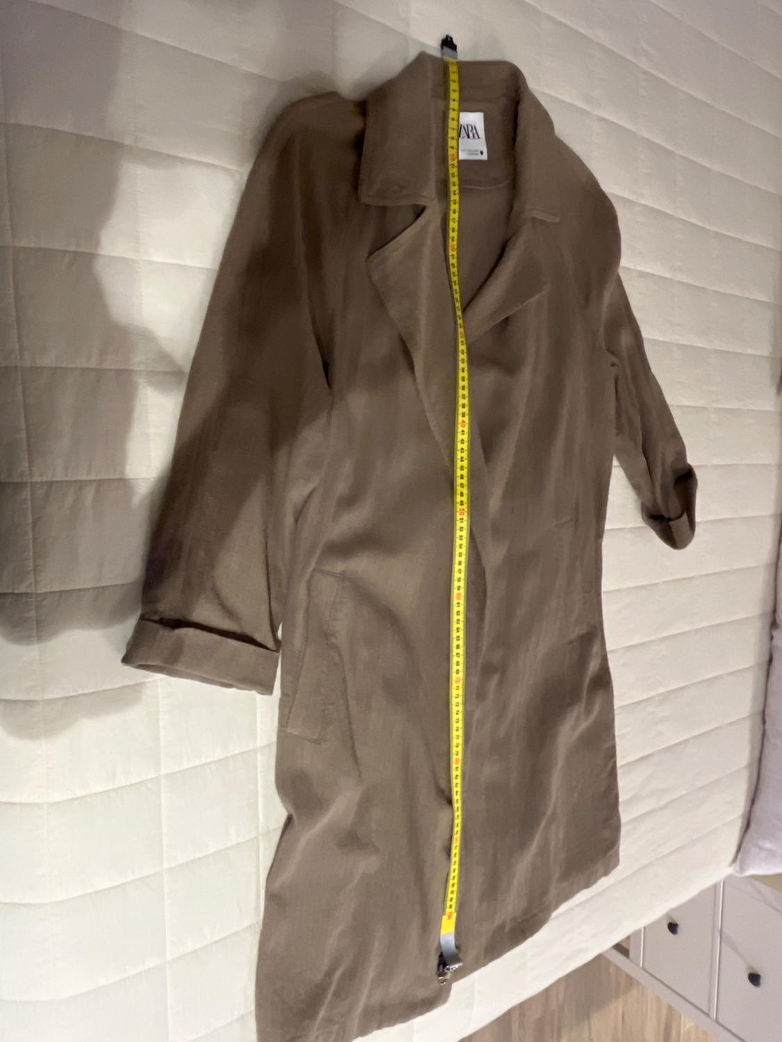 Light brown coat on a white bed, with a yellow tape measure measuring its length. Visible label on the collar.