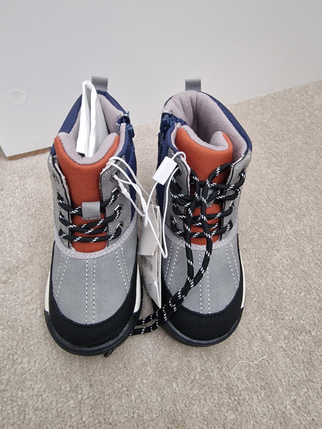 Children's winter boots in grey, blue and orange with black laces and labels, on beige carpet.
