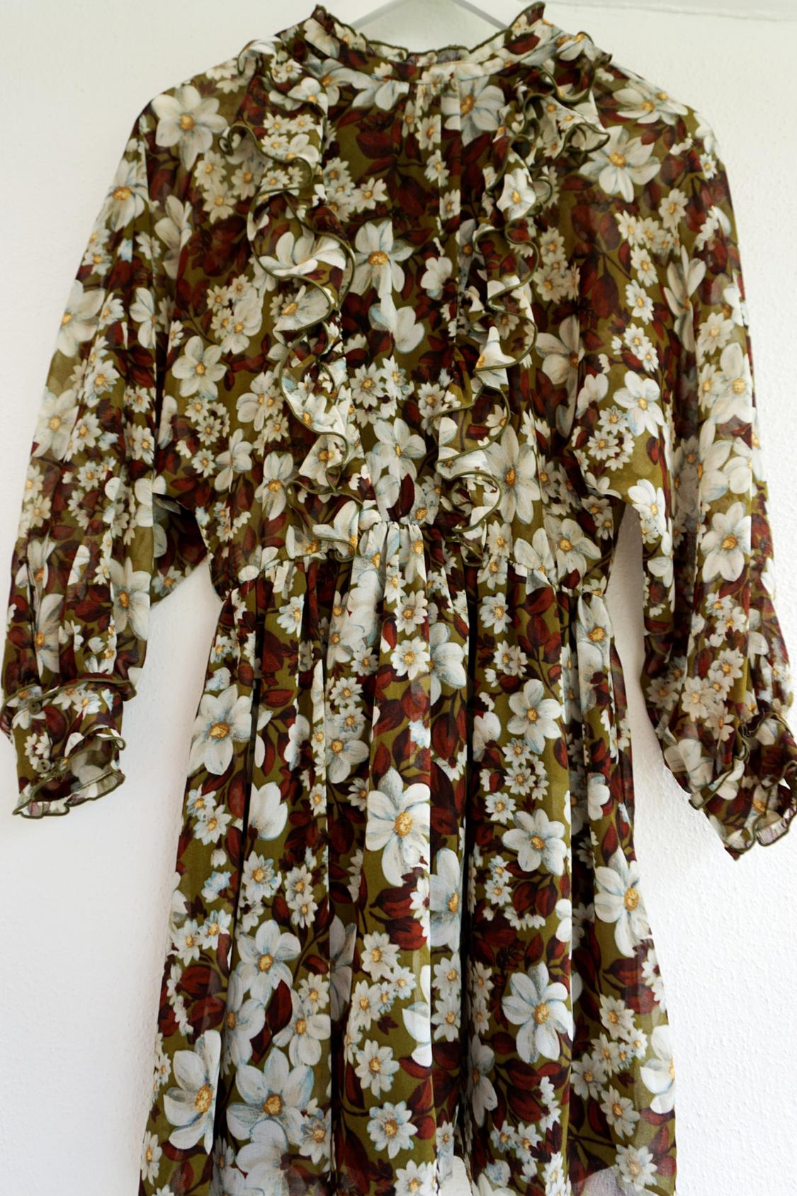 Floral dress with ruffles, olive green background and white and brown flowers. Long sleeves and a high neck, hanging on a white hanger.