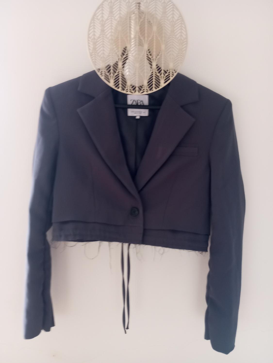 Black short jacket hanging on a hanger with a visible Zara label. White background and decorative lamp above.