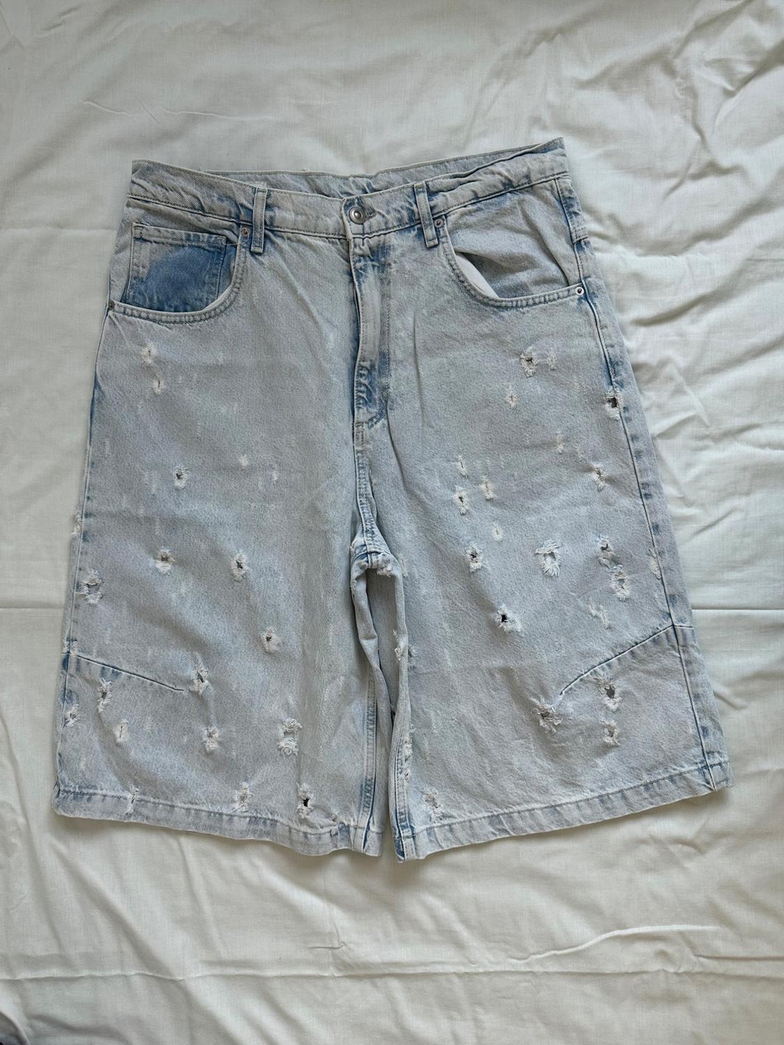 Light blue distressed denim Bermuda shorts, laid flat on a crumpled white fabric. Casual and relaxed style.