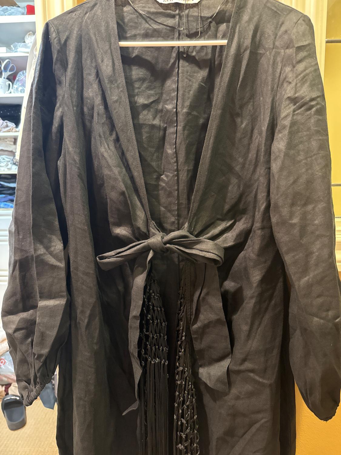 Black coat hanging on a hanger, with a tie at the waist and fringe details. Background with shelves and visible clothing.