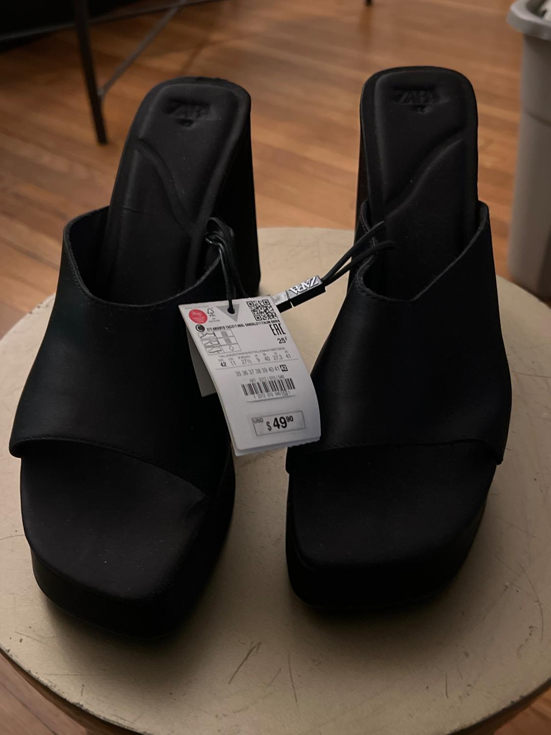 Black high-heeled sandals on a wooden table. Visible tag with a price of £49.90. Indoor setting.
