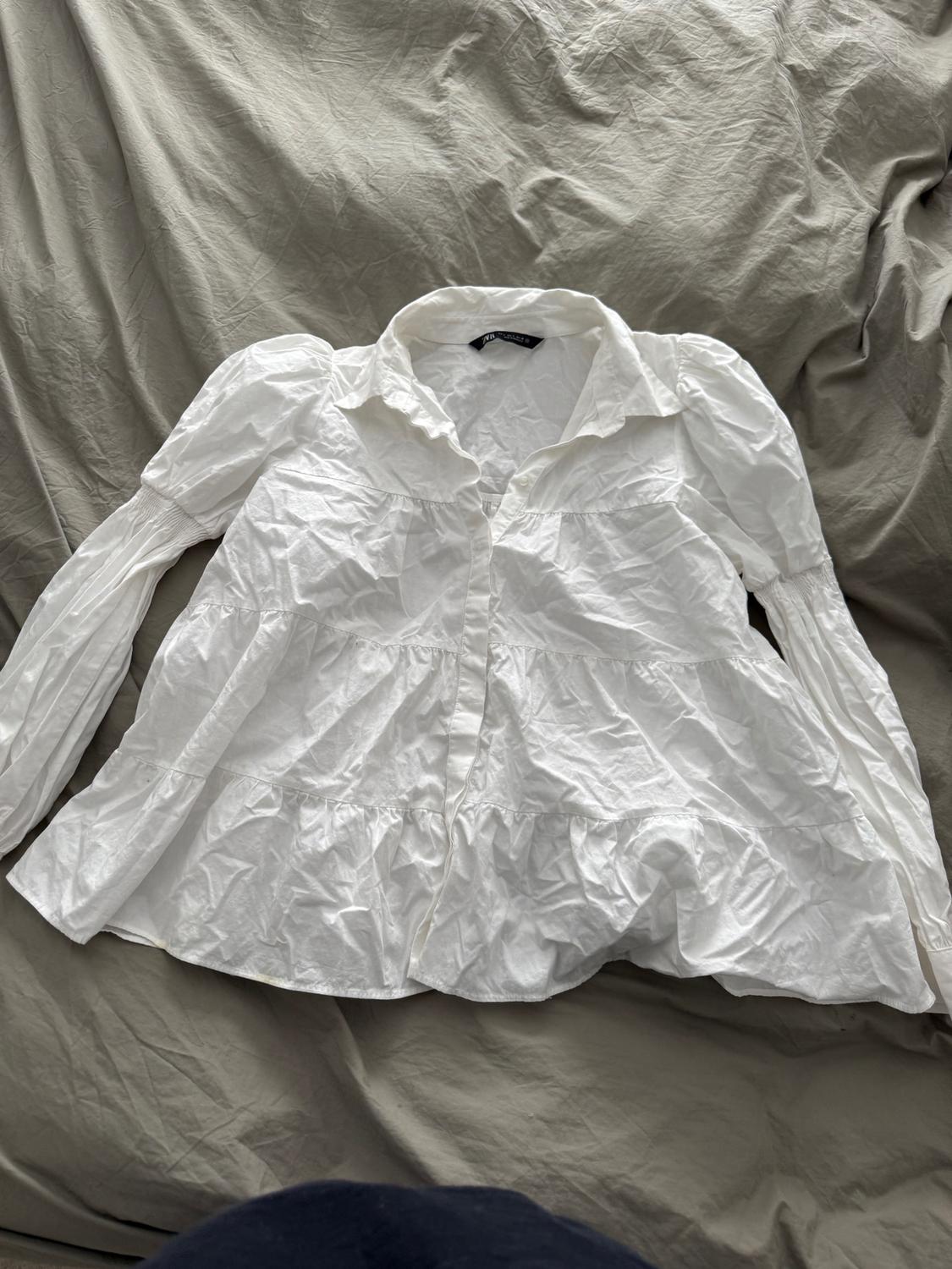 Wrinkled white long-sleeved collared shirt on a bed with gray sheets. Visible tag on the collar.