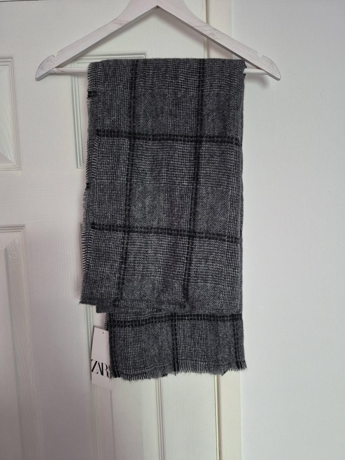 Grey checked scarf with black tassels hanging on a white hanger over a door. Label with the text ZARA.