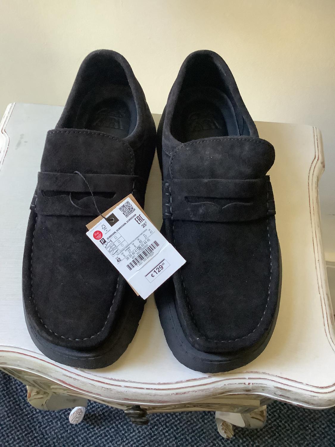 Black suede loafers on a white wooden stool. The left shoe has a tag with the text €129.00.