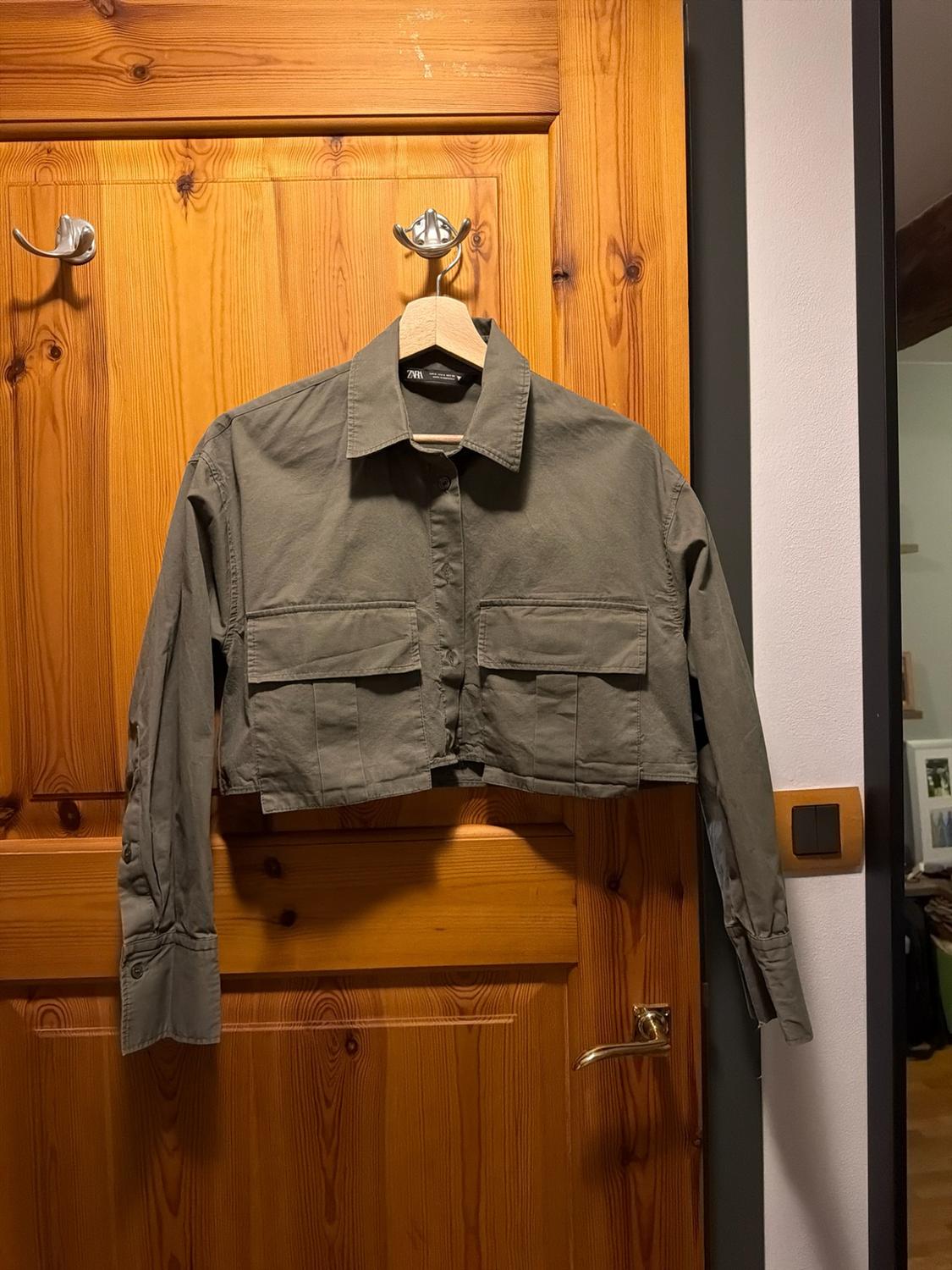 Short olive-green shirt with front pockets, hanging on a hanger over a wooden door. ZARA label.