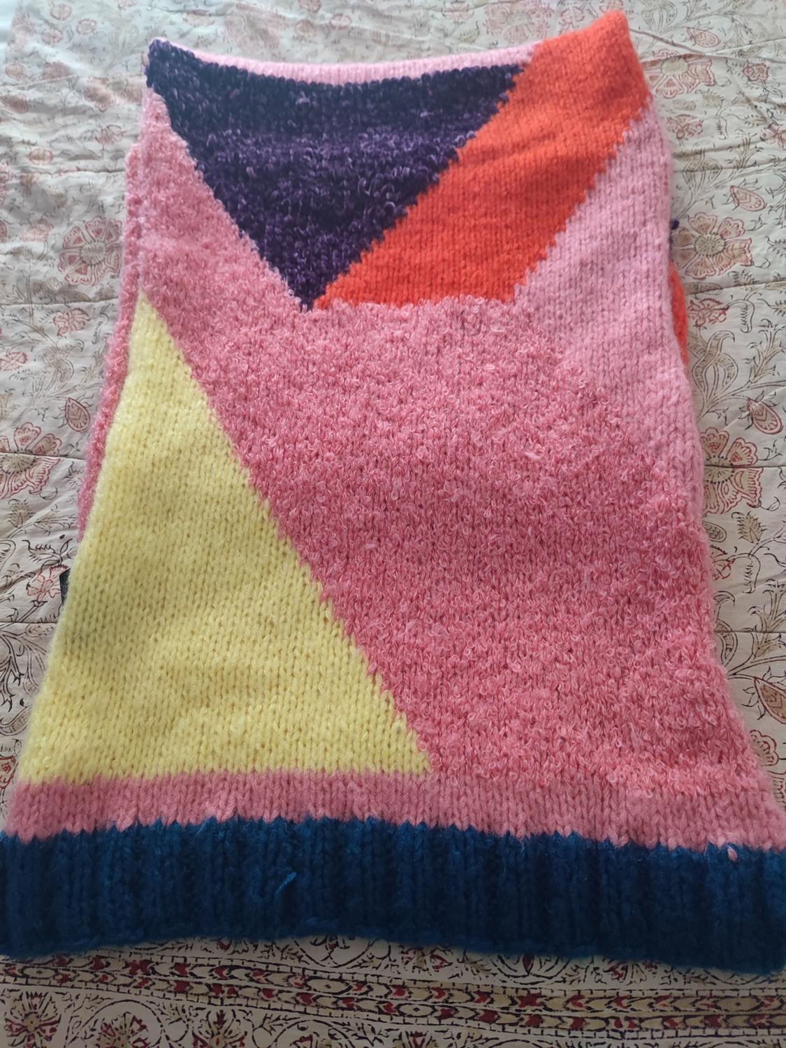 Knitted blanket with geometric design in pink, yellow, blue, orange, and purple on a floral patterned surface.