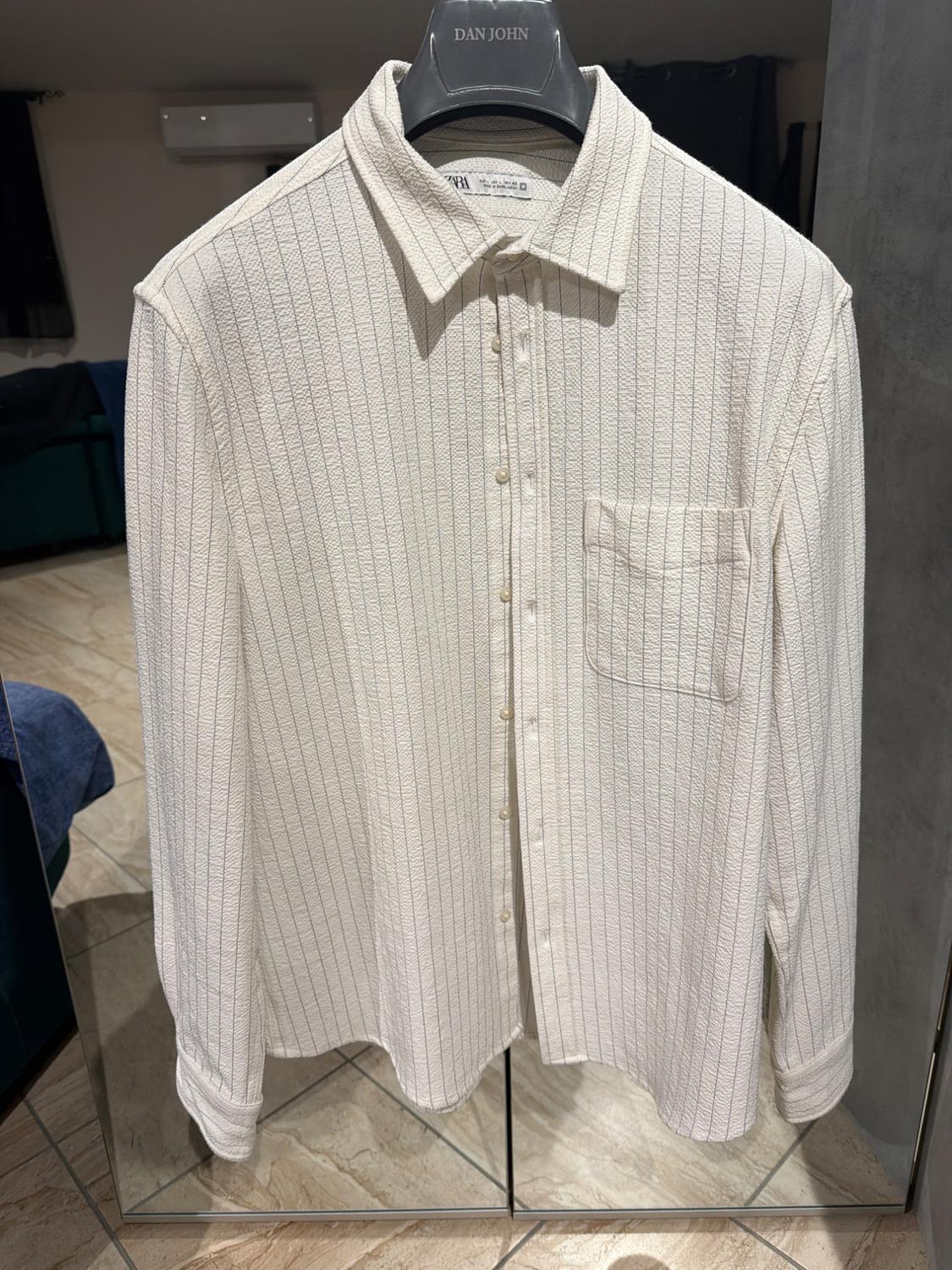 White shirt with vertical stripes hanging on a black hanger with the brand "DAN JOHN", reflected in a mirror in a room.
