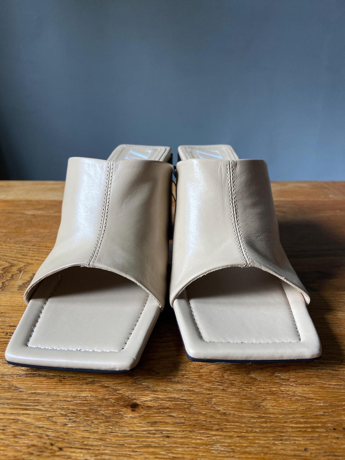Beige square-toe mule heeled sandals, presented on a wooden table
against a grey background.