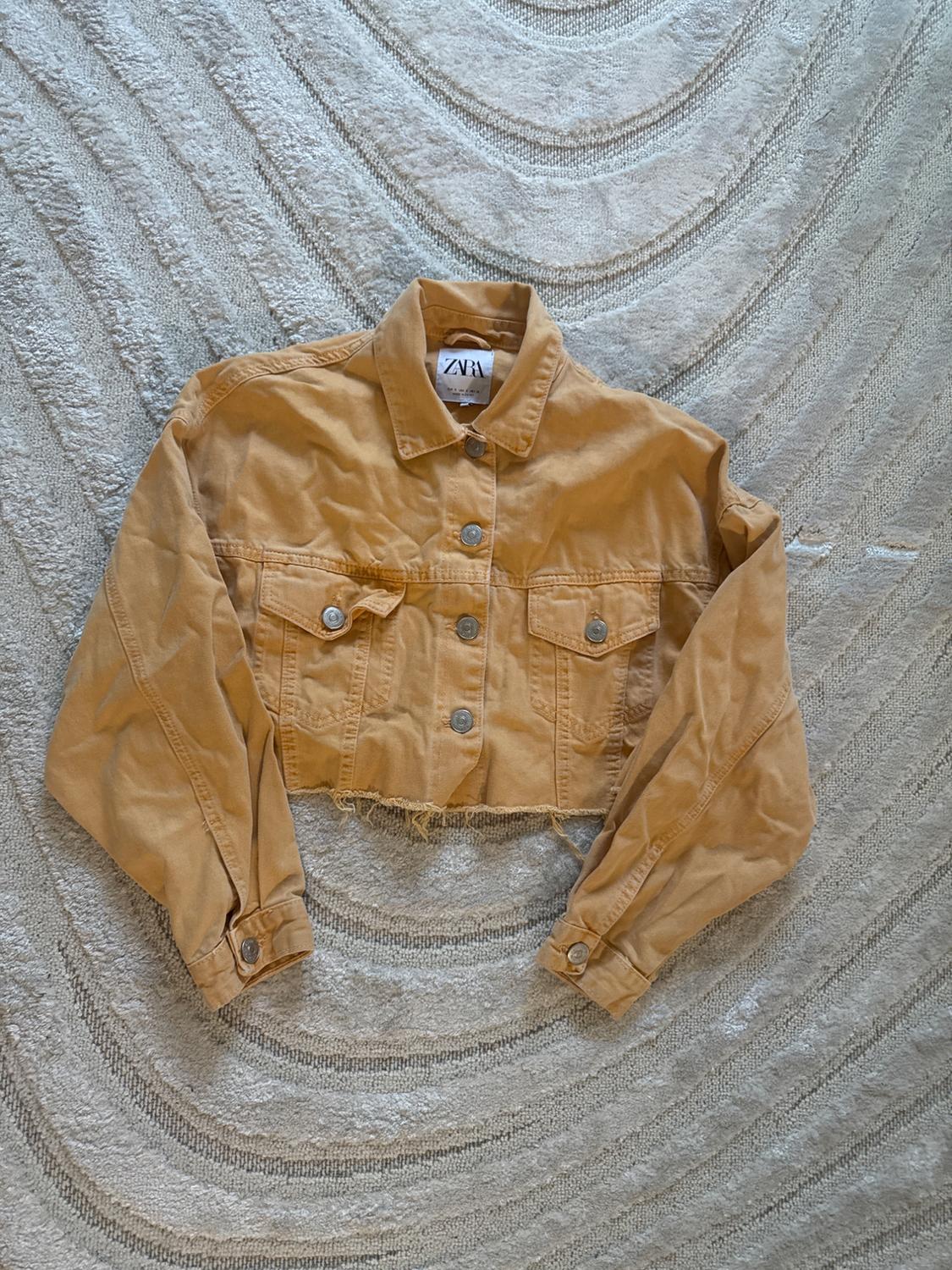 Short beige denim jacket with metallic buttons, on a textured rug in light tones. Visible tag: ZVÁ.