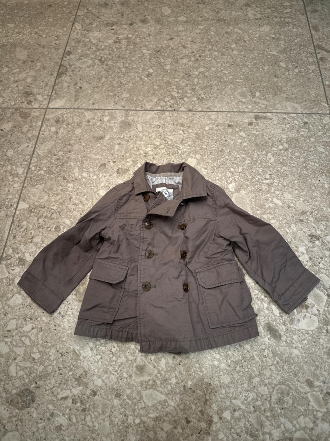 Grey children's coat with brown buttons and front pockets, on a grey tiled floor.