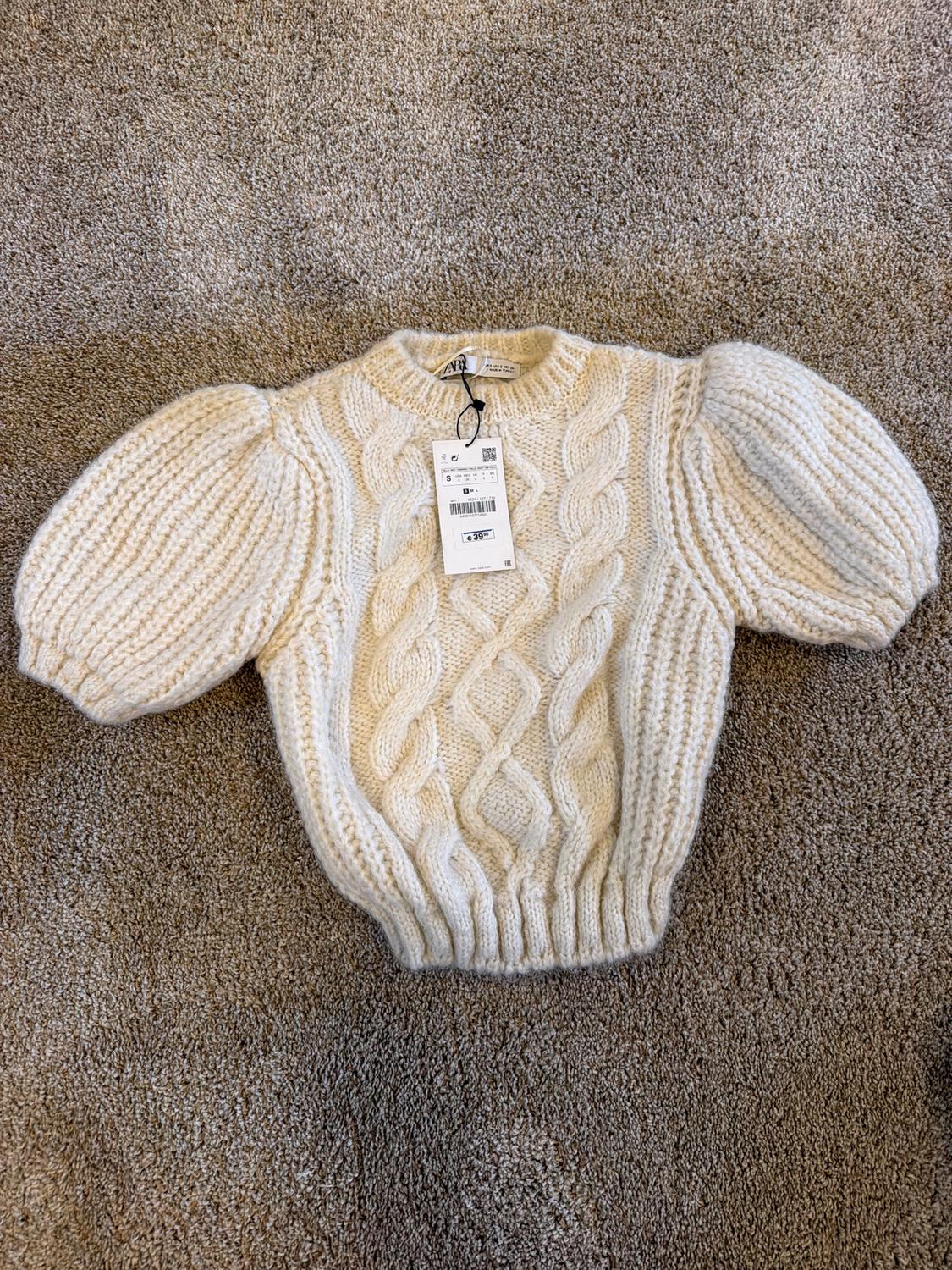 Chunky cream knit jumper with puff sleeves on a beige rug. Visible tag with price of €39.95.