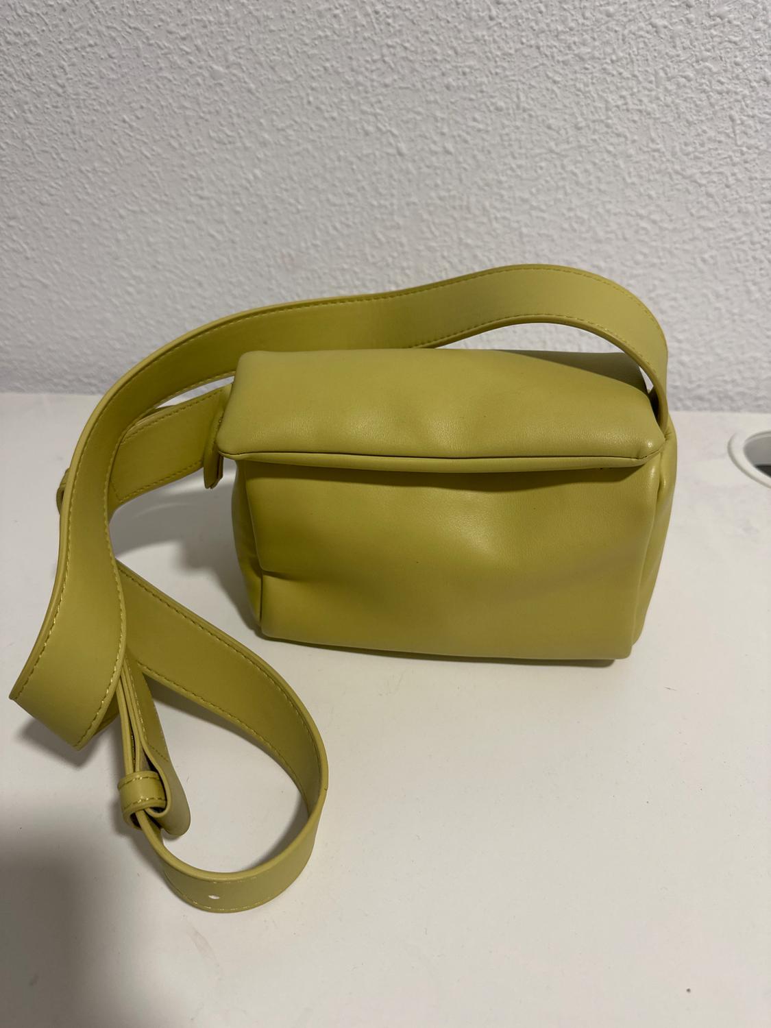 Light yellow handbag with an adjustable strap on a white surface, textured wall background.
