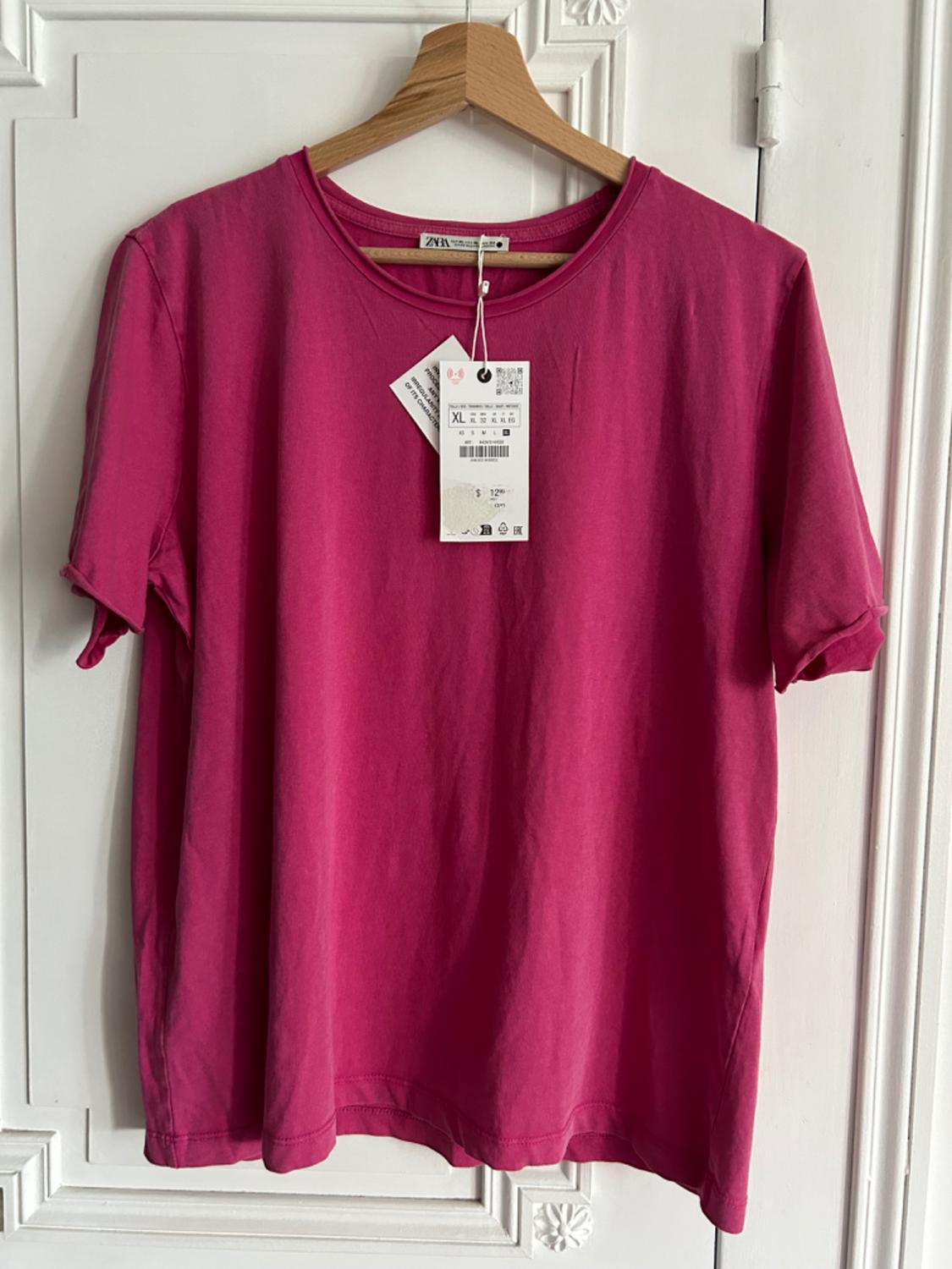 Pink t-shirt hanging on a wooden hanger, with visible labels indicating size XL and a price of €12.95, on a white door.