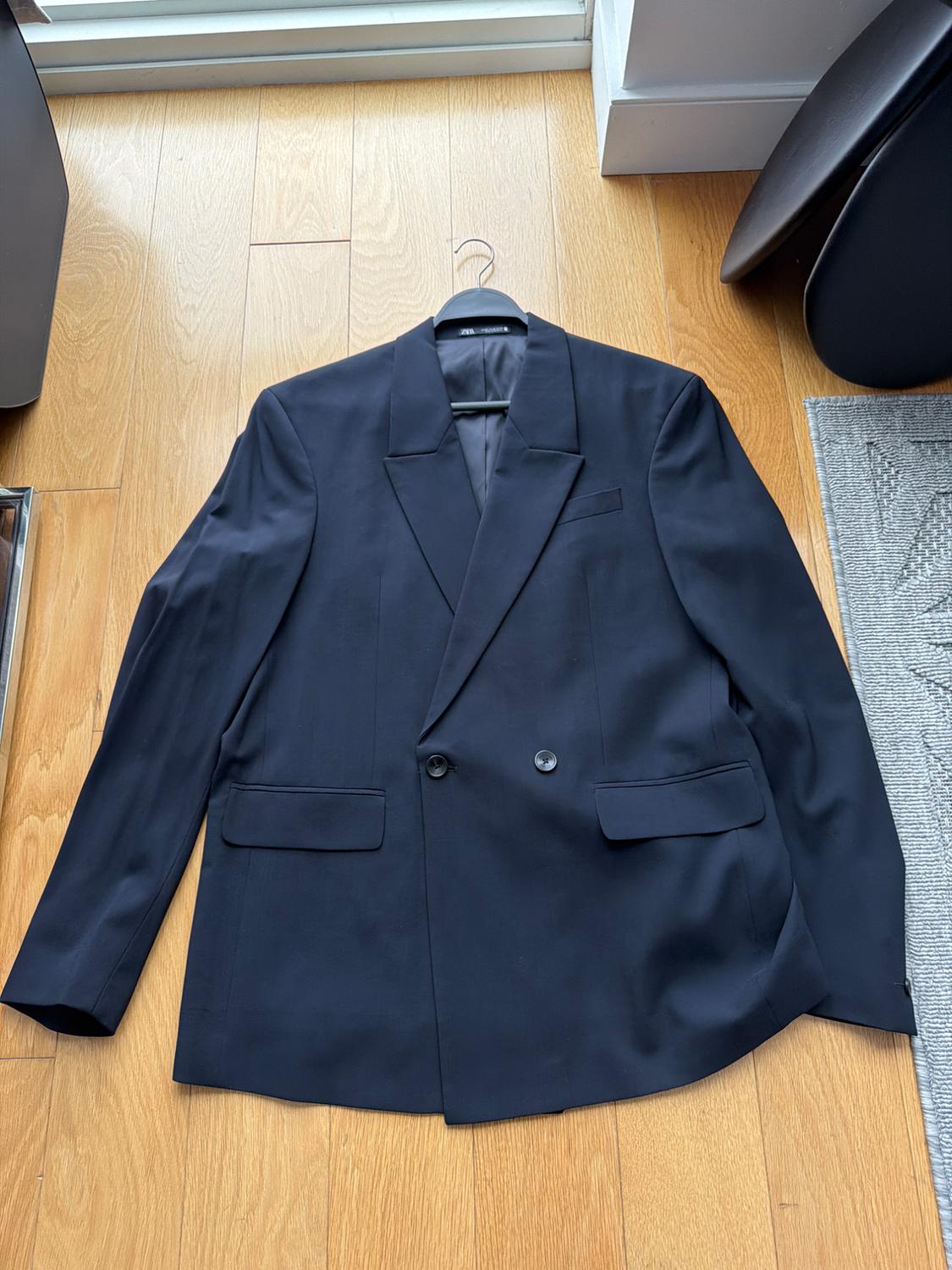 Black double-breasted suit jacket, hanging on a hanger over light wooden flooring, next to a gray rug.
