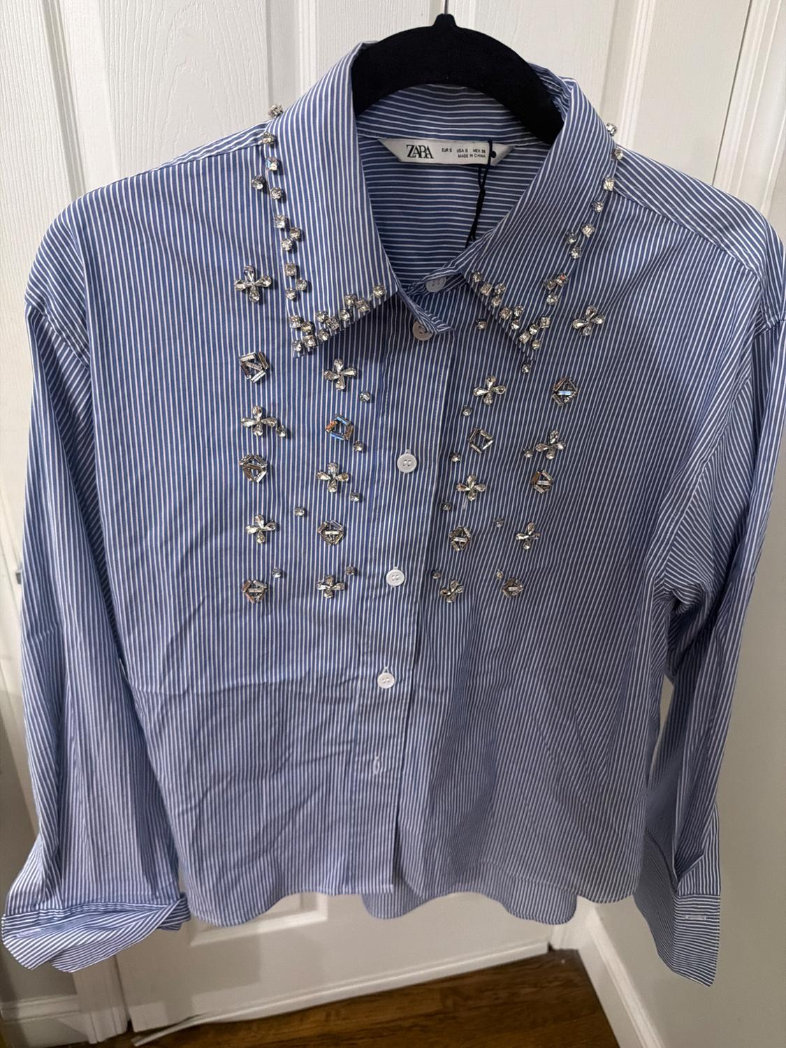 Blue and white striped shirt with pearl and crystal embellishments on the collar and chest, hanging on a black hanger.