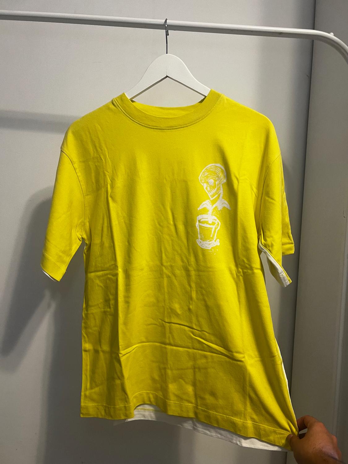 A yellow t-shirt is hanging on a white hanger, with a white skull and plant design on the chest. A hand is holding the edge.