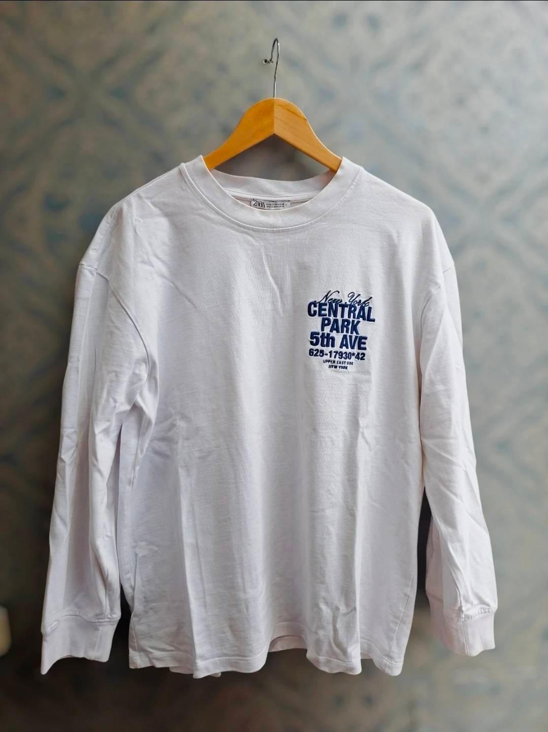 White long-sleeved t-shirt hanging on a wooden hanger. Blue text: "New York Central Park 5th Ave 625-17930*42 Upper East Side New York".