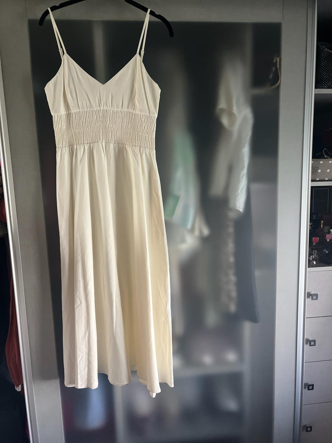 Long cream-coloured dress with thin straps and a gathered waist, hanging on a hanger in front of a translucent glass wardrobe.