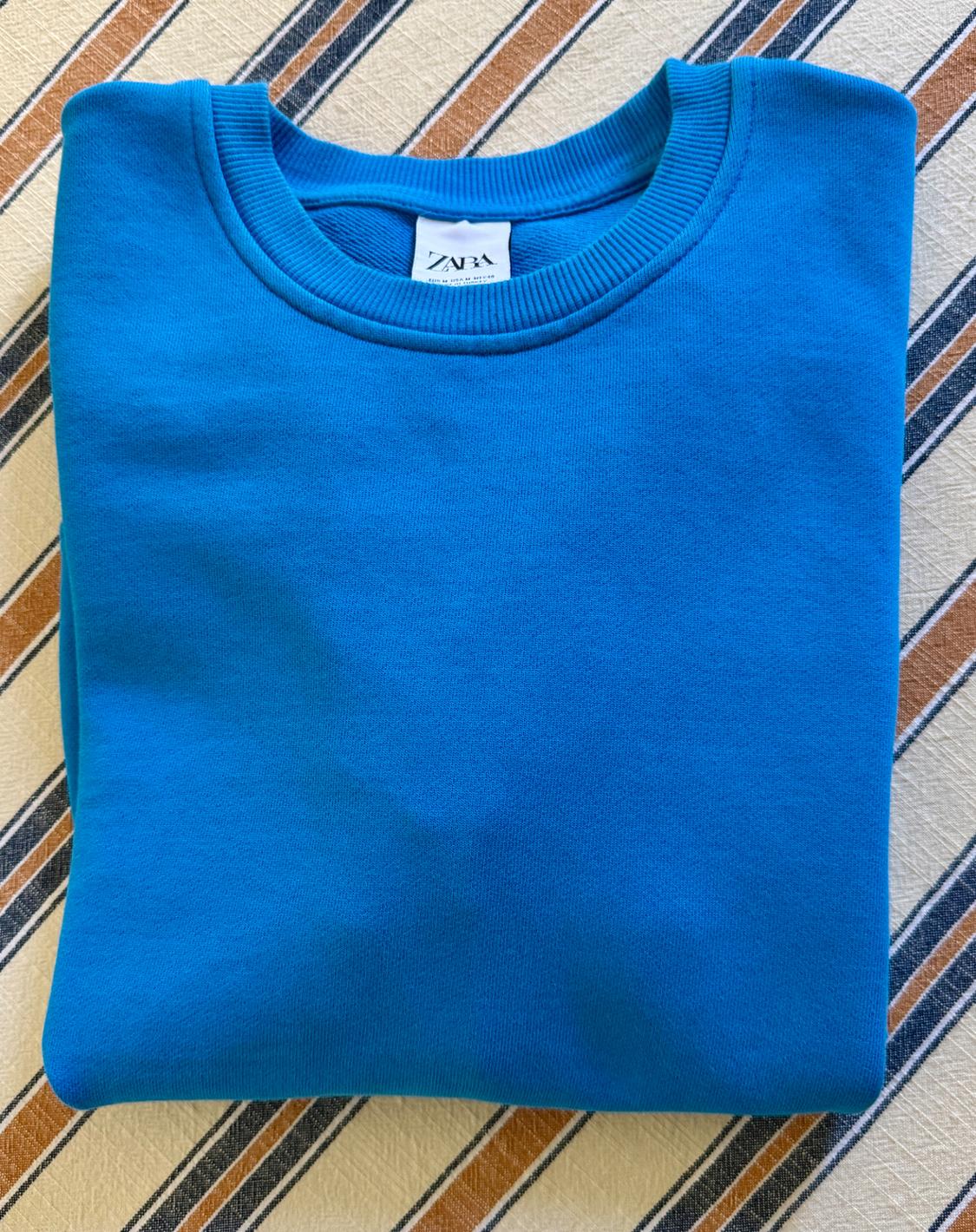 Blue sweatshirt folded on a fabric surface with diagonal stripes. The inner tag reads ZARA.
