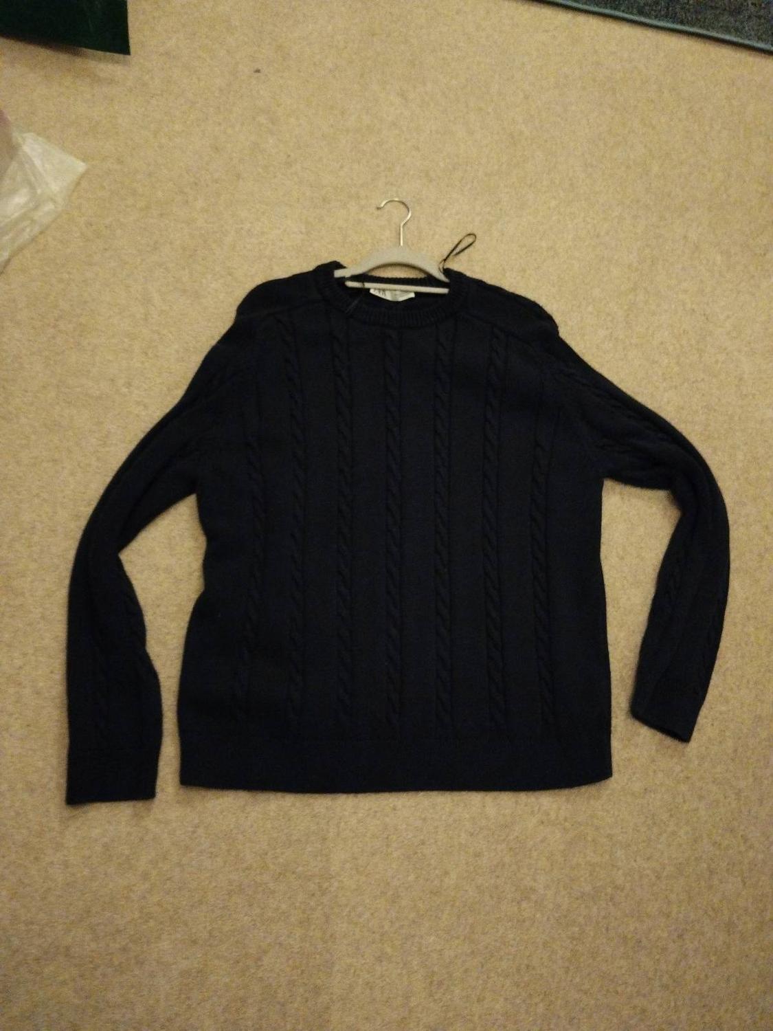 Dark cable-knit long-sleeved jumper, laid on a beige carpet with a white hanger peeking out from the neck.