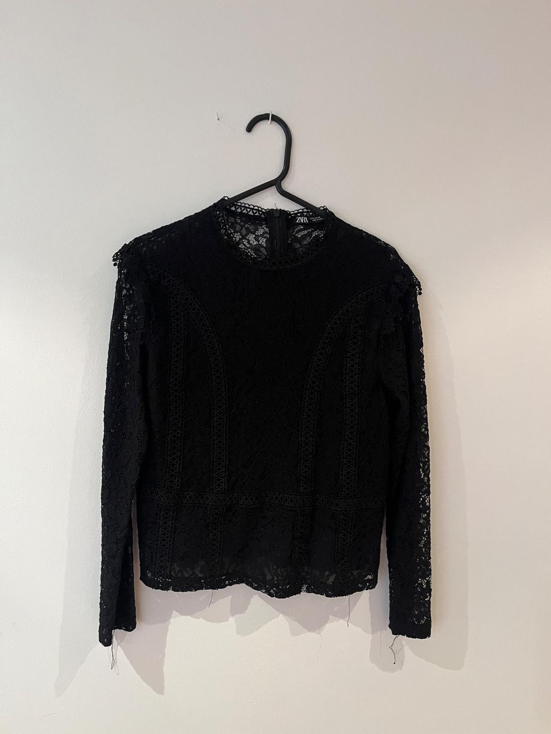 Black long-sleeved lace blouse hanging on a black hanger against a white wall.