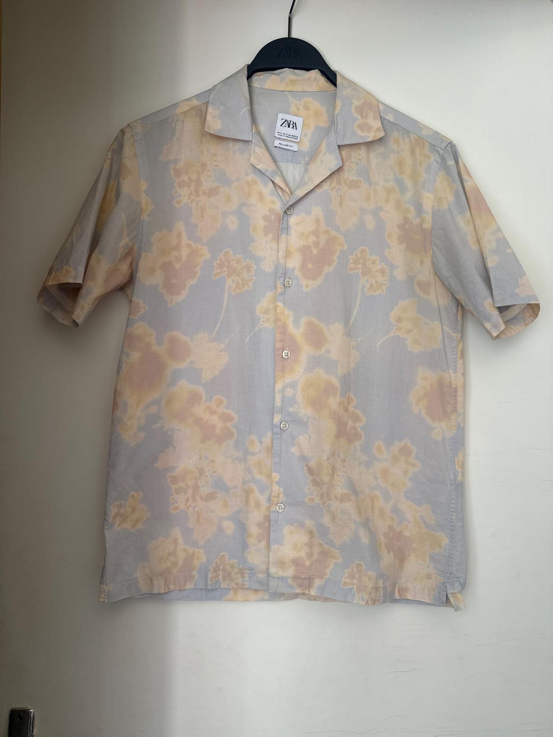 Short-sleeved shirt with faded floral print in yellow tones, hanging on a black hanger against a white background. Text: ZARA.
