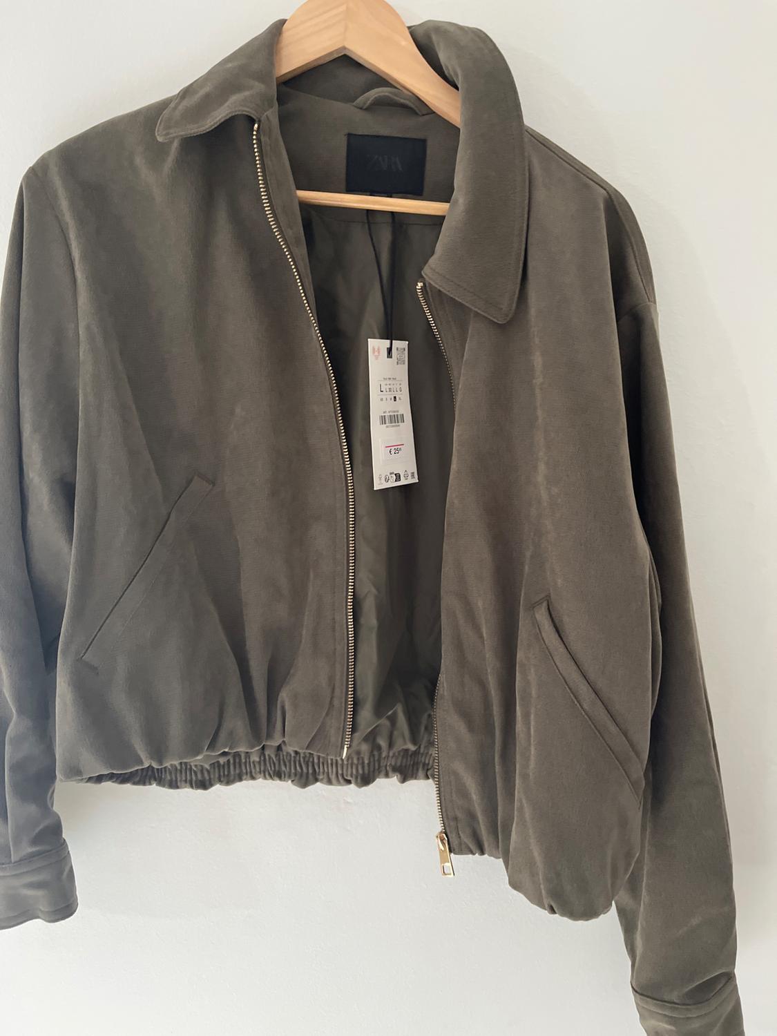 Olive green jacket with a front zip and elasticated hem, hanging on a wooden hanger against a white background. Text: ZARA, L.