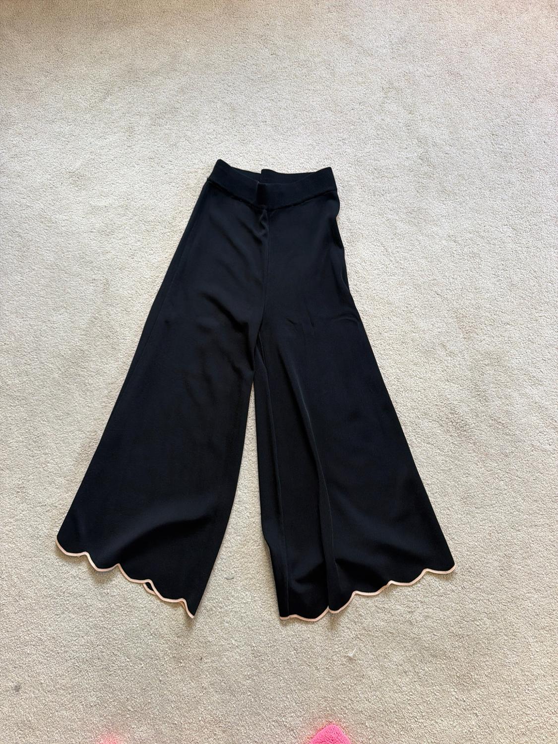 Black wide-leg trousers with a scalloped hem and light trim, laid out on a beige rug.