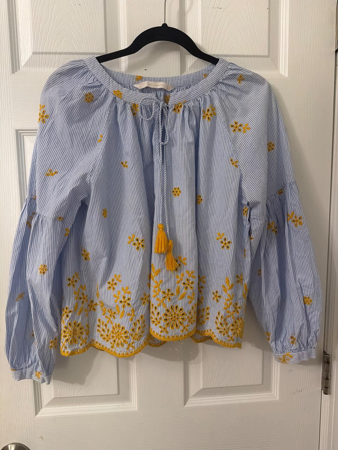 Blue and white striped blouse with yellow floral embroidery and a drawstring neckline, hanging on a black hanger in front of a white door.