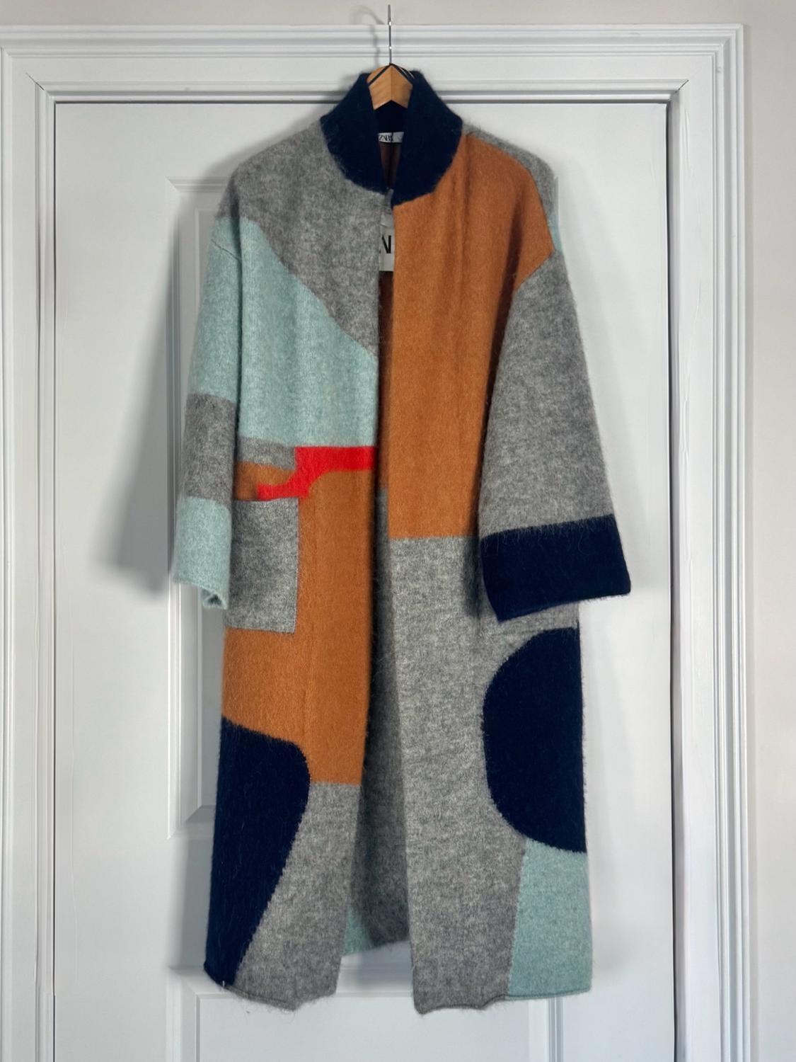 Longline coat with a soft texture and a design of geometric color blocks in gray, orange, light blue, and navy, hanging on a hook on a door.