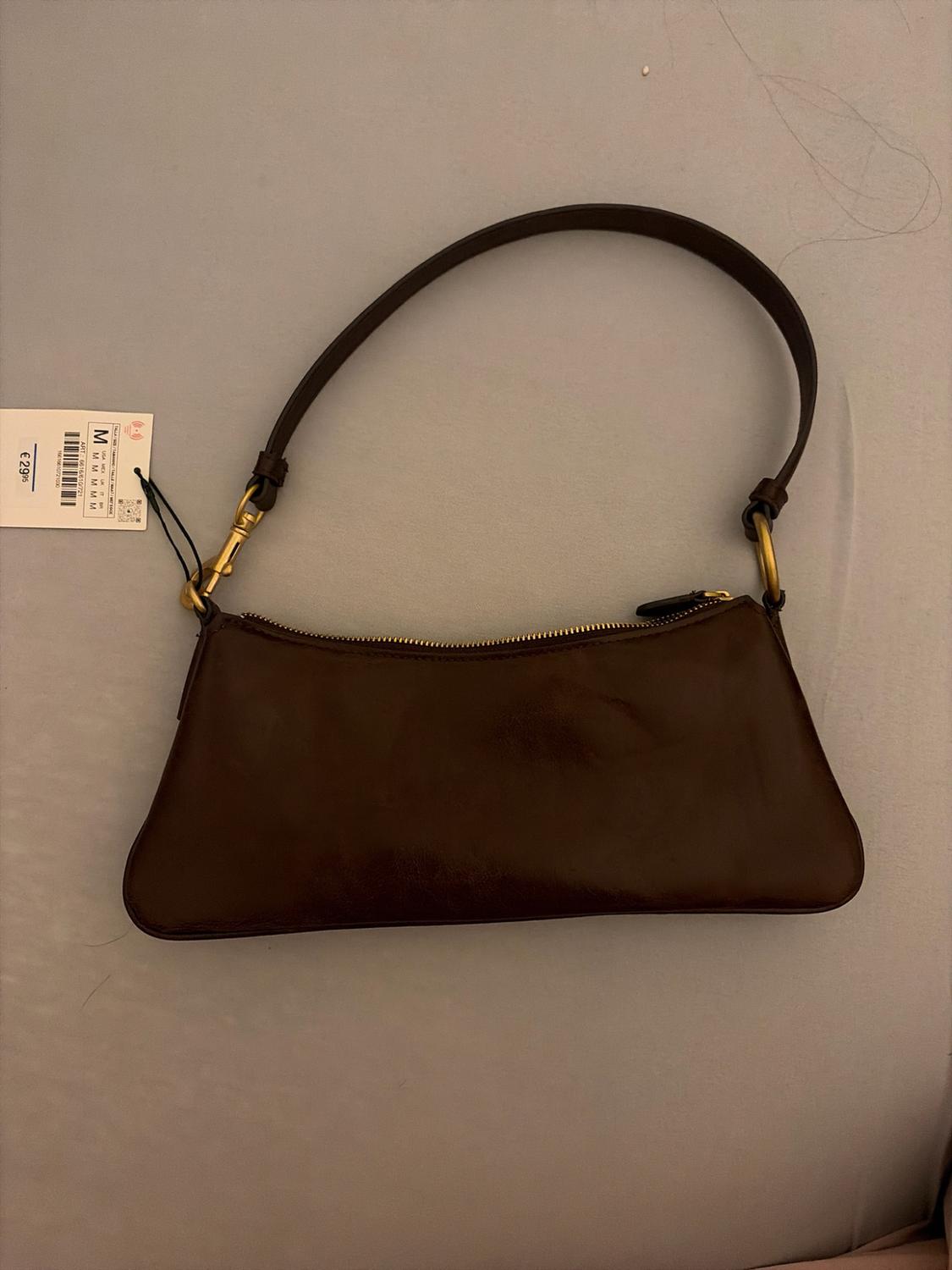 Dark brown shoulder bag with a handle and gold-coloured details on a fabric surface. Includes a tag with the text "M" and "€ 29.95".