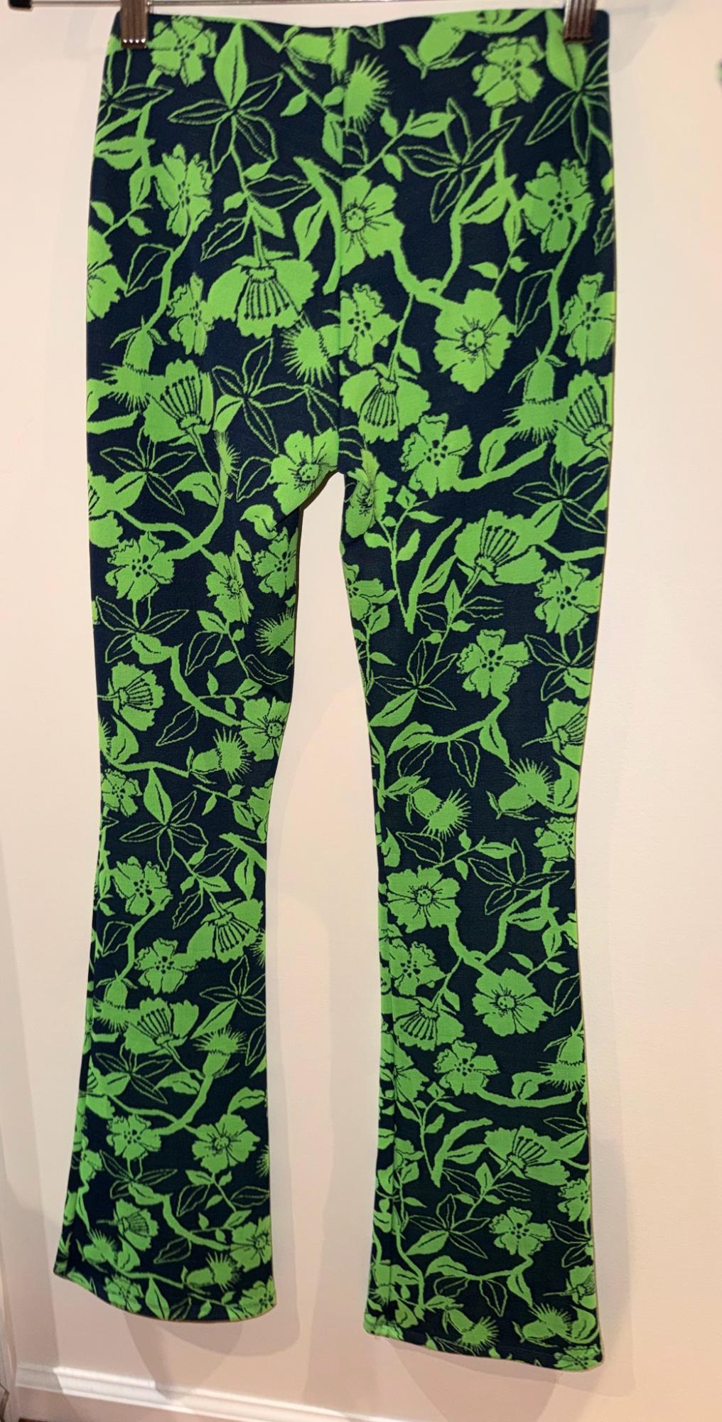 Flared trousers with a floral and green-leaf print on a dark background, hanging on a hanger against a white wall.