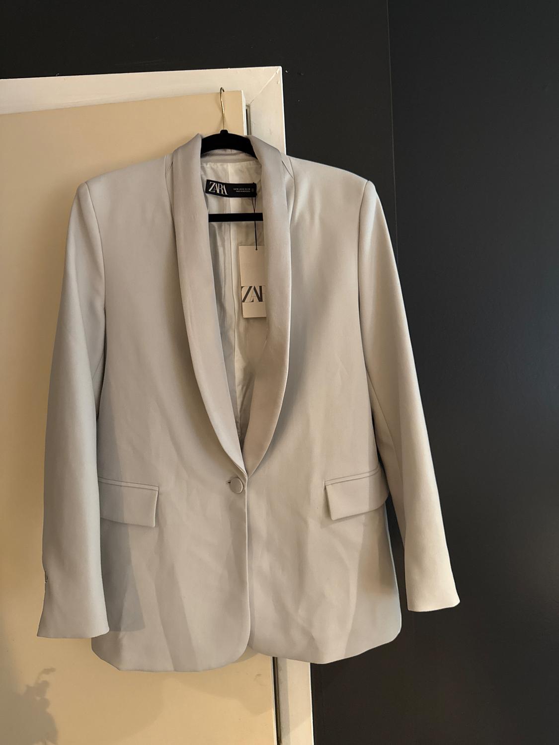 Light gray jacket hanging on a black hanger over a white door. Visible tag with Zara logo. Dark background.