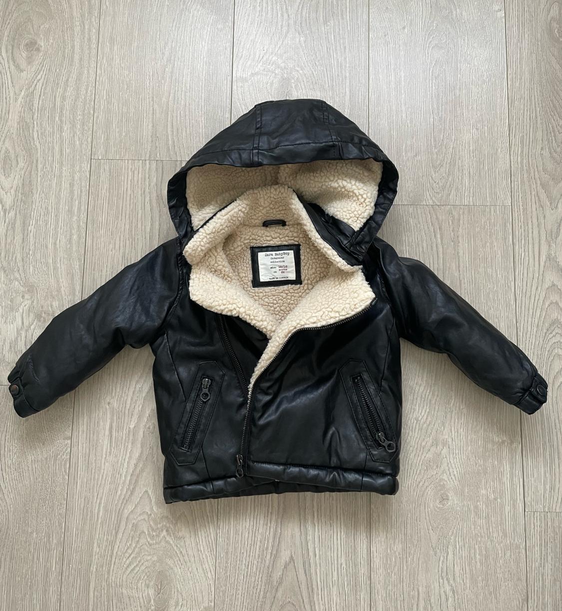Black boy's jacket with hood and white borg lining, on light wood floor. Label visible on the inside.