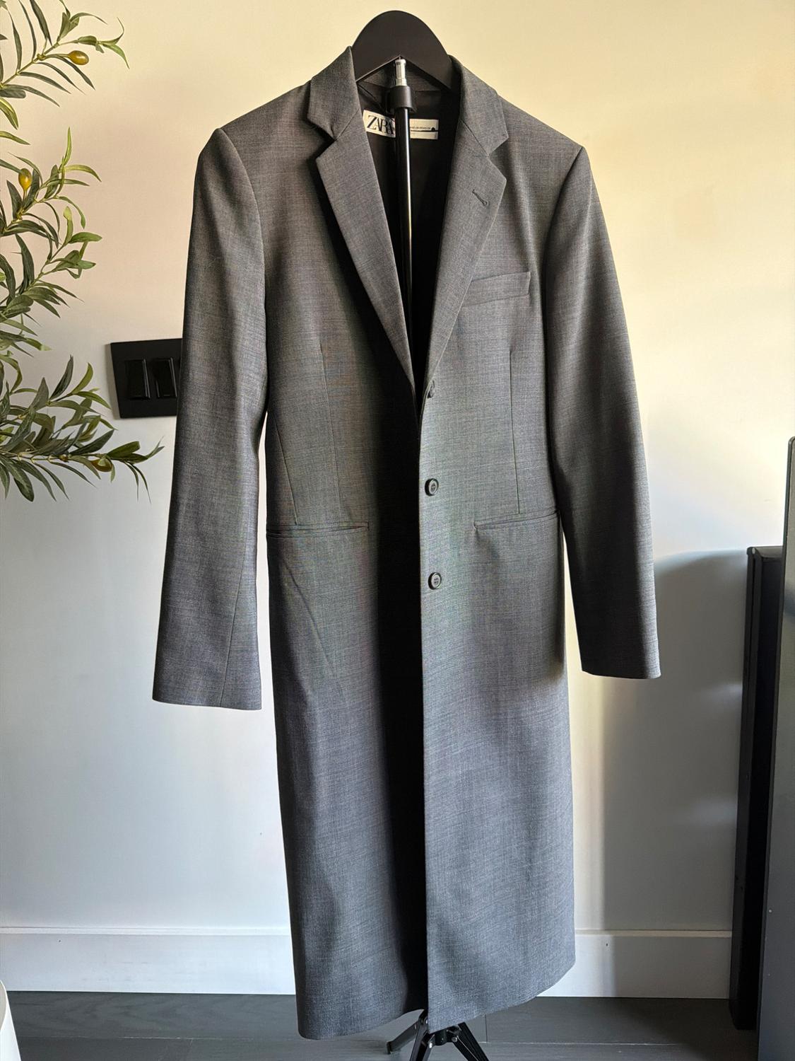 Long gray coat hanging on a black hanger, with a light wall background and a decorative plant on the left. Visible tag: ZVR.