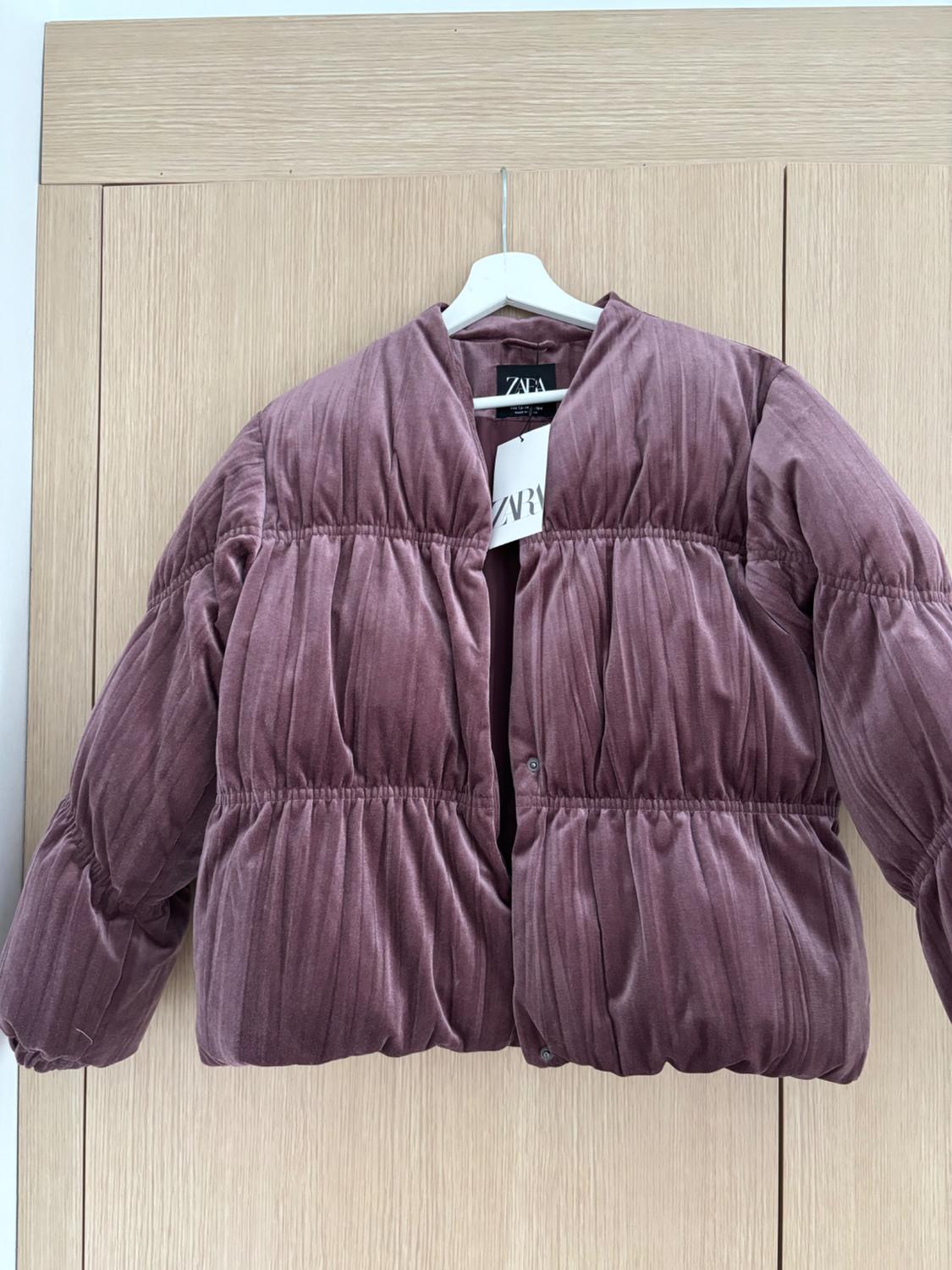 Mauve quilted jacket hung on a white hanger in front of a light wooden door. Visible tag with the text "ZARA".